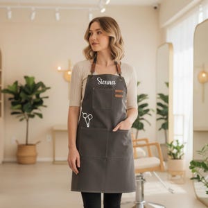 May include: A dark gray apron with brown leather straps and pockets. The apron has the name "Sienna" printed on the chest and a white graphic of scissors. The apron is designed for a salon or barber setting.