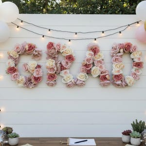 May include: A white wooden backdrop with the word "GRAD" spelled out in floral letters. The letters are made of pink and cream-colored roses. Balloons in gold, pink, and white are on either side. String lights hang above the letters.