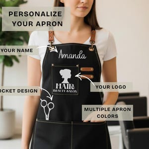 May include: Black apron with leather straps and pockets. The apron has the name "Amanda" in white script. A pocket features a silhouette of a woman's face and the words "Hair Beauty Salon". The apron is designed for personalization.