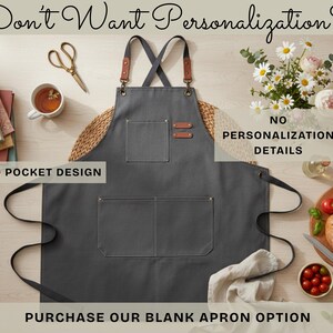 May include: A charcoal gray apron with two front pockets and leather accents. The apron has a cross-back design and is displayed with a bouquet of flowers, a cup of tea, and a loaf of bread. Text on the image reads: "Don't Want Personalization?" and "Purchase Our Blank Apron Option."