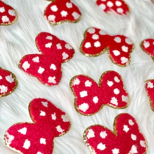May include: A collection of red and white polka dot bow-shaped fabric patches with gold trim. The patches are arranged on a white fuzzy surface.