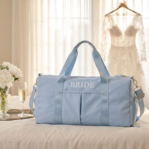 May include: A light blue duffel bag with the word "BRIDE" embroidered on the front. The bag has a shoulder strap and handles. A wedding dress hangs in the background, with a bouquet of white flowers and a glass of champagne on a silver tray.