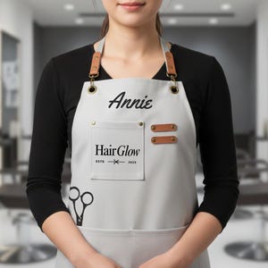 May include: A light gray apron with the name "Annie" printed in black script. The apron features a pocket with the text "Hair Glow" and a pair of scissors. The apron has brown leather straps and accents.