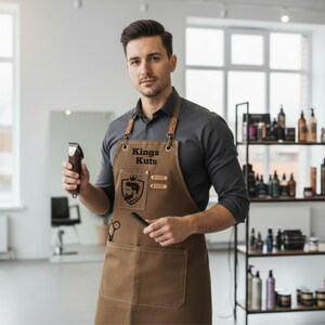 May include: A brown barber apron with the text "Kings Kuts" and a logo featuring a silhouette of a man's face. The apron has leather straps and pockets. The person is holding a hair clipper and a comb.