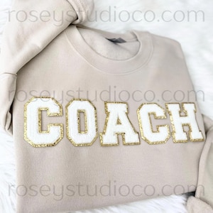 May include: A beige sweatshirt with the word "COACH" in white, gold-trimmed, block letters. The sweatshirt is folded on a white, fluffy surface. The brand name "roseystudioco.com" is printed on the sweatshirt.