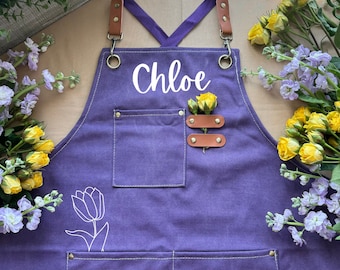 May include: A deep purple apron featuring the name "Chloe" in white script. It has brown leather details, a pocket, and a tulip outline. Yellow roses are attached to the apron.