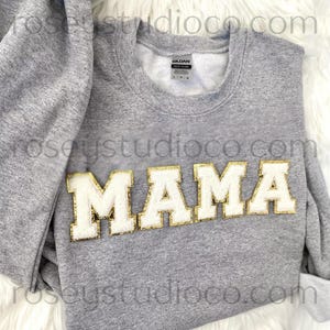 Könnte beinhalten: Graues Sweatshirt mit dem Wort "MAMA" in weißen Buchstaben mit goldfarbenem Rand. Das Sweatshirt ist gefaltet, wobei der Kragen und die Ärmel zu sehen sind. Das Markenetikett ist im Kragen sichtbar.