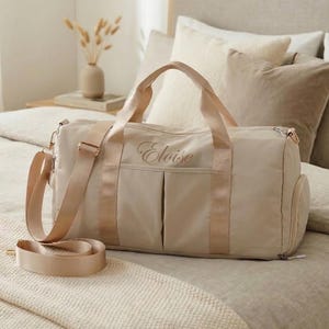 Peut inclure: Un sac de sport beige avec une bandoulière et des poignées. Le sac a un compartiment principal zippé et des poches latérales. Le nom "Eloise" est brodé sur le devant en écriture cursive. Le sac est sur un lit avec une literie beige.