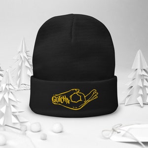 Puede incluir: Gorro negro con un gesto de mano bordado en oro y la palabra "GOTCHA" en el puño.