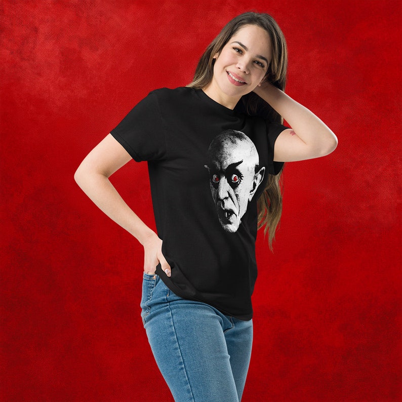 May include: A black t-shirt with a white and black graphic print of a man's face with red eyes. The man has a serious expression and is looking directly at the viewer.