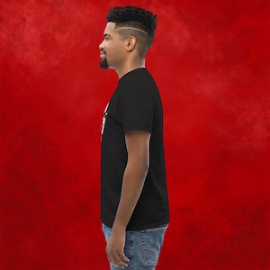 May include: A black t-shirt with a white graphic design. The design features a stylized image of a skull with a crown on top. The person wearing the shirt is standing in front of a red background.