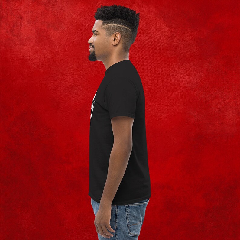 May include: A black t-shirt with a white graphic design. The design features a stylized image of a skull with a crown on top. The person wearing the shirt is standing in front of a red background.