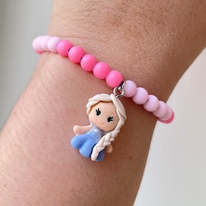 Könnte beinhalten: Ein rosa-weißes Perlenarmband mit einem Anhänger einer Zeichentrickfigur eines Mädchens mit blonden Haaren, das ein blaues Kleid trägt.