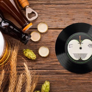 May include: A black vinyl record with a green apple design and the text "The Beatles -  Sgt. Pepper's Lonely Hearts Club Band" on the label. The record is on a wooden surface with beer bottles, bottle openers, and bottle caps.