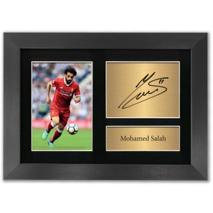 May include: A framed photo of a soccer player in a red and white uniform with a black background. The player is holding a soccer ball and is running on a green field. The photo is next to a gold plaque with a black signature and the name "Mohamed Salah" printed below.