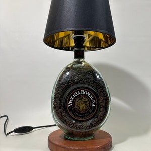 May include: A table lamp made from a clear glass bottle filled with coffee beans. The bottle has a label that reads "Vecchia Romagna" with a picture of a person. The lamp has a black lampshade with a gold lining and a wooden base.