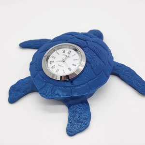 May include: A blue ceramic turtle clock with a silver face and Roman numeral markings. The clock reads "CAMRY QUARTZ".