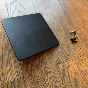 May include: A square, matte black plate with rounded corners, accompanied by two small silver-toned metal T-nuts and two small black screws. The plate features a smooth, solid surface and is presented alongside the necessary hardware for assembly.