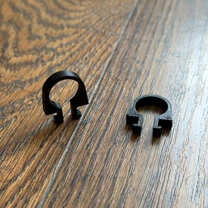 May include: Two black, circular, 3D-printed clips with a C-shaped opening at the bottom. One clip stands upright, while the other lies flat on its side. Both objects feature a textured surface and a small, notched design at the base of the opening.