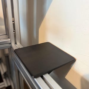 May include: A flat, square, matte black shelf component attached to a silver aluminium extrusion frame. The shelf features a smooth, solid surface and is mounted horizontally, extending outward from the vertical metal support structure.