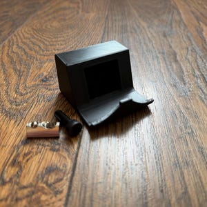 May include: A black 3D-printed plastic mount with a square opening and a curved front lip, accompanied by a small silver metal cylindrical fastener and a black screw, designed for securing or holding a specific component in place.