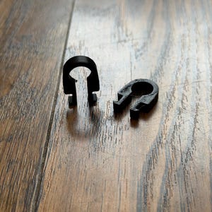 May include: Two small, black, U-shaped plastic clips with a circular opening at the top and two downward-facing prongs at the base. One clip stands upright while the other lies flat on its side, both featuring a textured, matte finish.