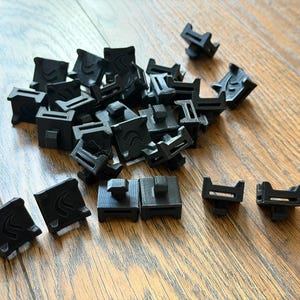 May include: A collection of small, black, square-shaped plastic clips. Each clip features a raised, curved design on the top surface and a protruding tab on the underside. The components are arranged in a loose pile with several pieces laid out flat in the foreground, showing their uniform construction and matte finish.