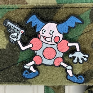 May include: A blue and pink cartoon character with a gun, holding a gun in its right hand. The character is wearing blue shoes and has a big smile. The character is on a green camouflage background.
