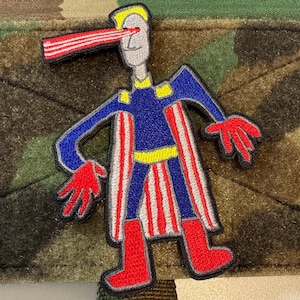 May include: Embroidered patch featuring a superhero-like figure with a blue and red costume. The figure has a yellow belt and accents, red boots and gloves, and a red beam shooting from its eye. The patch is on a camouflage background.