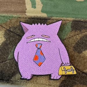May include: A purple embroidered patch featuring a cartoon character with a grumpy expression, wearing a blue tie with orange polka dots, and carrying a yellow briefcase. The patch is set against a camouflage background.