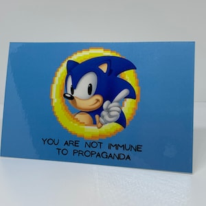 May include: A blue and yellow card with a cartoon image of Sonic the Hedgehog. The text on the card reads "You are not immune to propaganda."