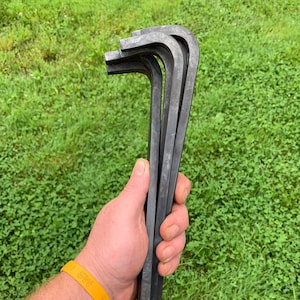 May include: Three black metal tent pegs, approximately 30 cm long, with a slight bend at the top, held in a hand.