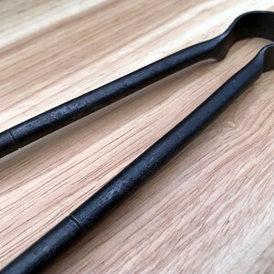 May include: Black metal tongs with a rounded handle. The tongs are on a light brown wooden surface.