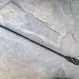 Könnte beinhalten: Ein langer, handgeschmiedeter Kaminbesteck-Schürhaken aus dunkelgrauem Eisen mit einer spitzen, hakenförmigen Spitze an einem Ende und einem dekorativen, gedrehten Metallgriff, der in einer kreisförmigen Schlaufe zum Aufhängen endet.