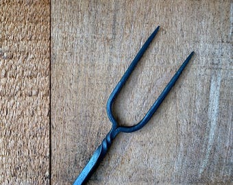 May include: A hand-forged black metal two-pronged fork featuring a twisted handle design. The utensil has a dark, matte finish with a long, slender neck that splits into two sharp, pointed tines, showcasing a textured, industrial appearance.