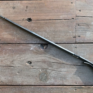 May include: A long, hand-forged black metal fire poker with a pointed hook at one end and a looped handle at the other. The shaft features a square cross-section, and both ends are decorated with a twisted metal detail near the hook and handle.