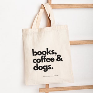 May include: A white canvas tote bag with black text that reads "books, coffee & dogs." The text is centered on the bag and is in a bold, sans-serif font. The bag has a brown leather handle and is hanging from a wooden ladder.
