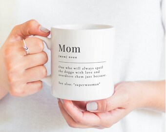 May include: White ceramic mug with the definition of "Mom" printed in black. The definition includes the phrase "One who will always spoil the doggo with love and overdress them just because." The mug is held by a person.
