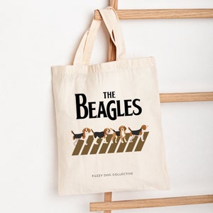 May include: A white canvas tote bag with black text that reads "The Beagles" and a graphic of five beagle dogs walking across a brown striped sidewalk. The text "Fuzzy Dog Collective" is printed in black at the bottom of the bag.