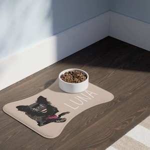 May include: A tan dog food mat with a black and white illustration of a dog with the name "LUNA" printed on it. A white ceramic dog bowl with brown dog food is on the mat.