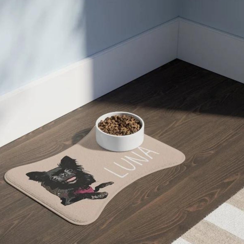 May include: A tan dog food mat with a black and white illustration of a dog with the name "LUNA" printed on it. A white ceramic dog bowl with brown dog food is on the mat.