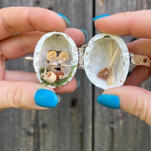 Può includere: Un diorama in miniatura all'interno di un guscio di noce, con un minuscolo topo con una tazza e una scopa in miniatura. Il guscio è aperto, rivelando l'interno dettagliato. Il diorama è tenuto tra due mani su uno sfondo di legno.