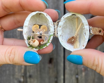 May include: A miniature diorama inside a walnut shell, featuring a tiny mouse with a cup and a miniature broom. The shell is open, revealing the detailed interior. The diorama is held between two hands against a wooden background.