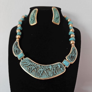 May include: A turquoise and gold-tone necklace and earring set. The necklace features a central pendant with giraffe carvings, turquoise accents, and gold-tone metal. The earrings have a matching design. The necklace is strung with gold-tone beads and turquoise square beads.