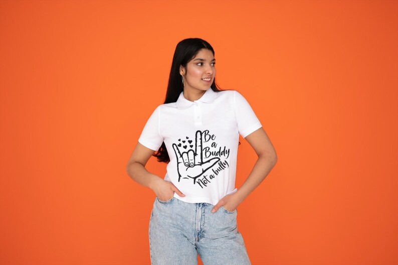 May include: White polo shirt with the text "Be a Buddy Not a Bully" in black. The shirt features a hand gesture graphic with hearts. The model is wearing light blue jeans against an orange background.