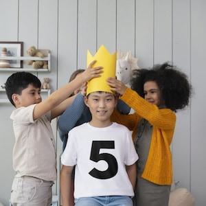 May include: A group of children placing a yellow paper crown on a smiling child wearing a white t-shirt with a large black number 5. The children are in a room with light gray walls and a shelf with toys.