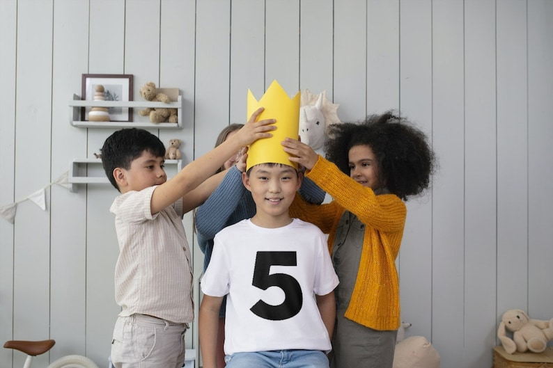 May include: A group of children placing a yellow paper crown on a smiling child wearing a white t-shirt with a large black number 5. The children are in a room with light gray walls and a shelf with toys.