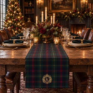 May include: A rectangular table runner featuring a dark green, navy blue, red, and gold plaid pattern. The fabric is centered on a wooden table and displays an embroidered gold crest with the letter K centered at the bottom edge.