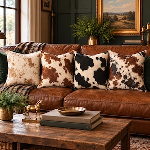 May include: Four square throw pillows arranged on a brown leather sofa. The pillows feature cowhide patterns in varying shades of white, brown, and black. The two outer pillows have light brown and white spots, while the inner two have dark brown and black patches on white backgrounds.