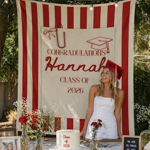 Può includere: Scena di festa di laurea con uno striscione che recita "CONGRATULAZIONI Hannah CLASSE DEL 2026". Una donna in abito bianco e cappello di laurea sorride. Una torta con "Classe del 2026" è su un tavolo con fiori e un cartello che dice "SIAMO COSÌ ORGOGLIOSI DI TE!"